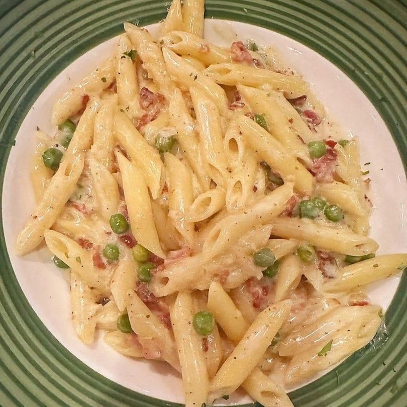 Creamy penne pasta with peas and bacon bits on a white plate with green stripes.